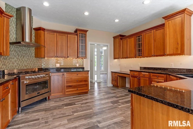 kitchen featuring stainless steel range with gas stovetop, wall chimney exhaust hood, dark stone countertops, and light wood-style floors