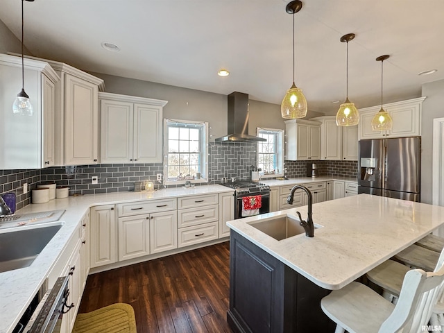 kitchen featuring an island with sink, appliances with stainless steel finishes, wall chimney range hood, pendant lighting, and a sink