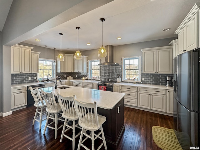 kitchen featuring an island with sink, gas range, wall chimney exhaust hood, light countertops, and refrigerator with ice dispenser