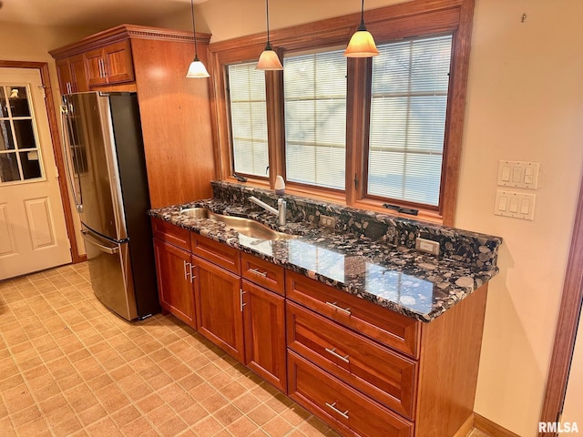 kitchen featuring a sink, freestanding refrigerator, brown cabinetry, dark stone countertops, and decorative light fixtures