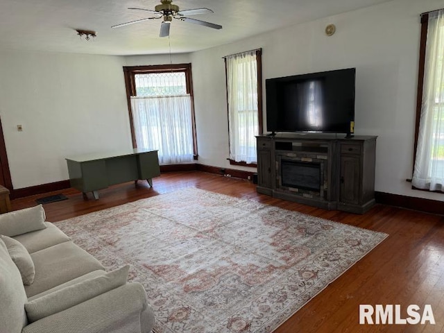 living area featuring a glass covered fireplace, ceiling fan, baseboards, and wood finished floors