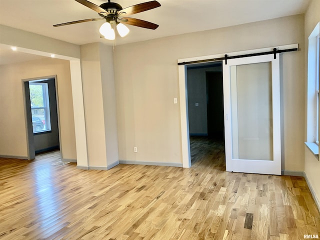 empty room with light wood finished floors, ceiling fan, a barn door, and baseboards