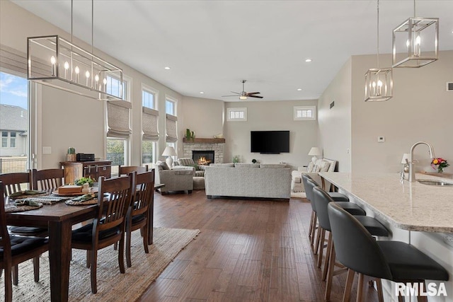 dining area with recessed lighting, a towering ceiling, dark wood-type flooring, a stone fireplace, and ceiling fan with notable chandelier