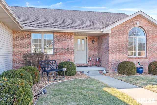 doorway to property with brick siding, a yard, and roof with shingles