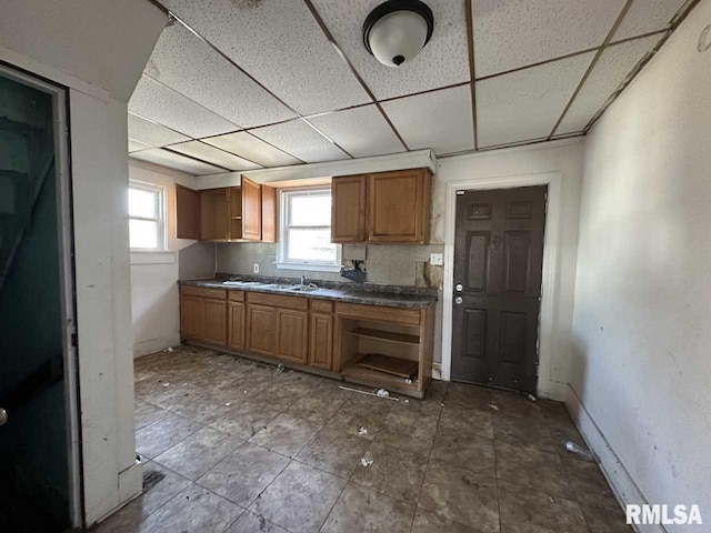 kitchen with dark countertops, brown cabinetry, a sink, a drop ceiling, and baseboards