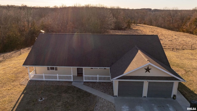 view of front facade featuring a shingled roof, covered porch, an attached garage, driveway, and a forest view