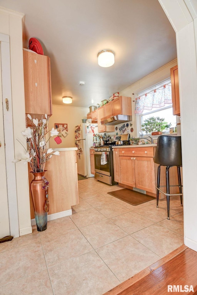 kitchen with light tile patterned floors, gas stove, freestanding refrigerator, light brown cabinetry, and under cabinet range hood