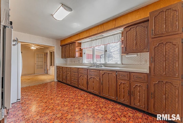 kitchen with brick patterned floor, freestanding refrigerator, brown cabinetry, wallpapered walls, and light countertops