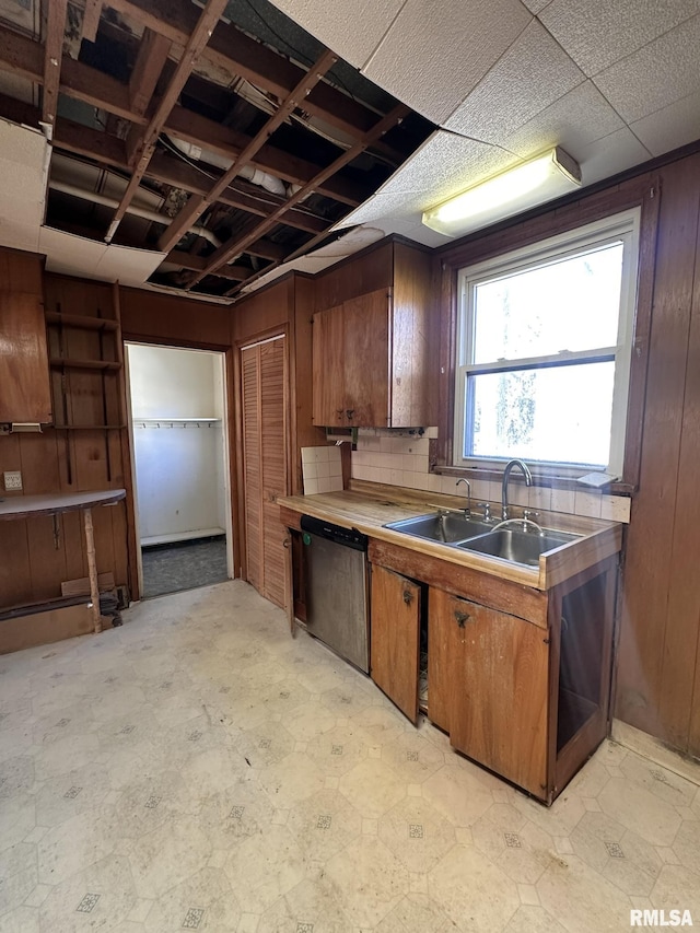 kitchen featuring a sink, stainless steel dishwasher, wood walls, brown cabinetry, and light floors