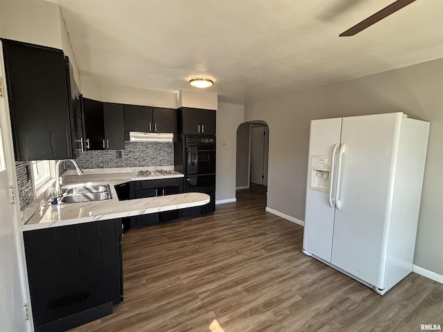 kitchen with dark cabinetry, arched walkways, a sink, under cabinet range hood, and white fridge with ice dispenser