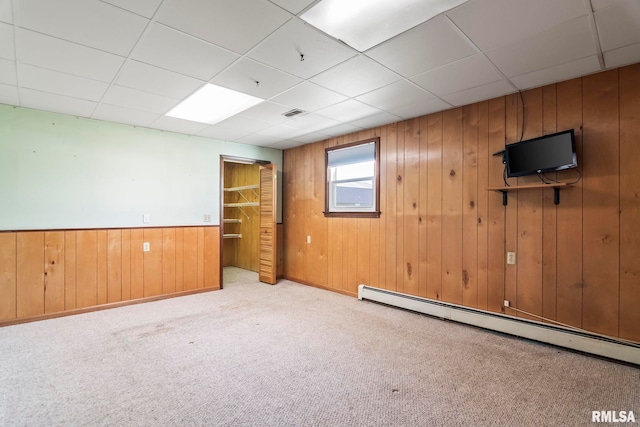 carpeted empty room with a drop ceiling, visible vents, wooden walls, and a baseboard radiator