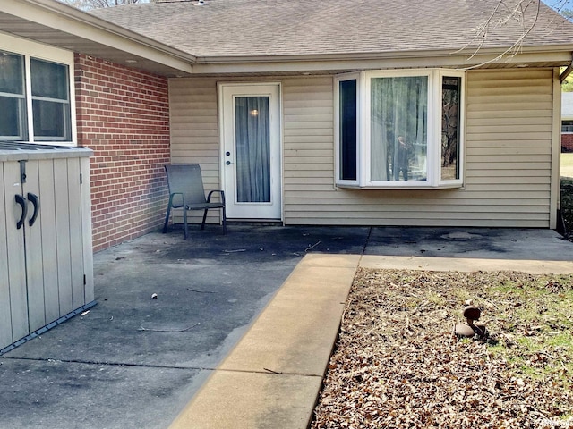 view of exterior entry with a patio area, brick siding, and a shingled roof