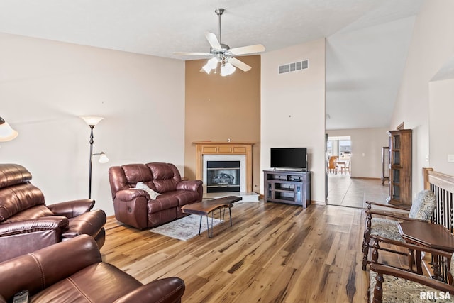 living room featuring light wood finished floors, visible vents, a glass covered fireplace, high vaulted ceiling, and a ceiling fan