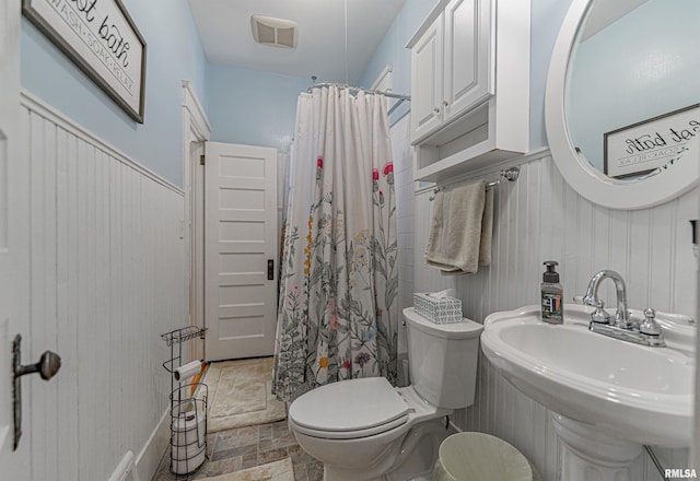 full bathroom featuring visible vents, toilet, a sink, stone finish flooring, and wainscoting