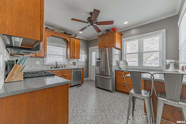 kitchen with brown cabinetry, light floors, ceiling fan, ornamental molding, and stainless steel appliances