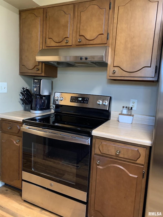 kitchen featuring under cabinet range hood, brown cabinetry, light wood finished floors, stainless steel electric range oven, and light countertops