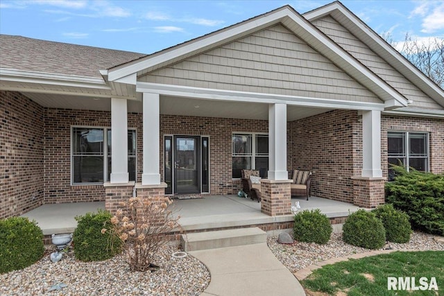 doorway to property featuring brick siding, a porch, and roof with shingles