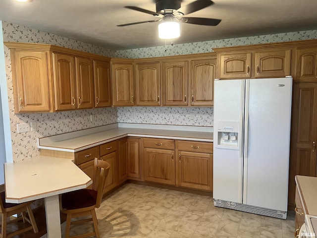 kitchen featuring light countertops, ceiling fan, wallpapered walls, brown cabinets, and white fridge with ice dispenser