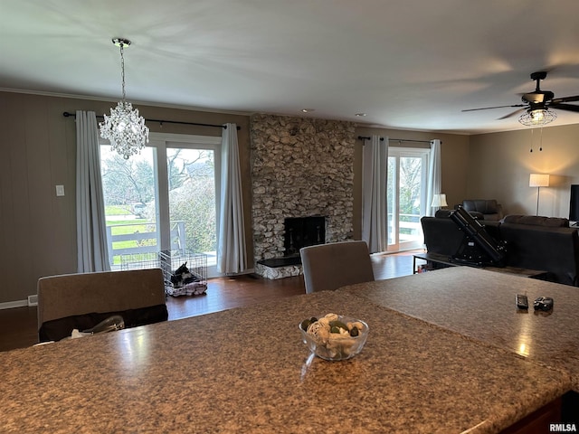 dining room with dark wood-style floors, a stone fireplace, and ceiling fan with notable chandelier