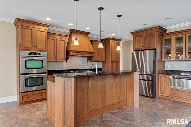 kitchen featuring appliances with stainless steel finishes, premium range hood, a kitchen island with sink, brown cabinets, and a warming drawer