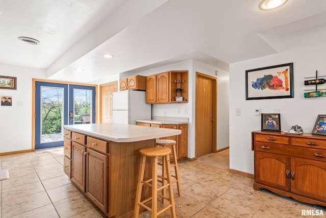 kitchen with baseboards, freestanding refrigerator, a kitchen bar, brown cabinetry, and light countertops