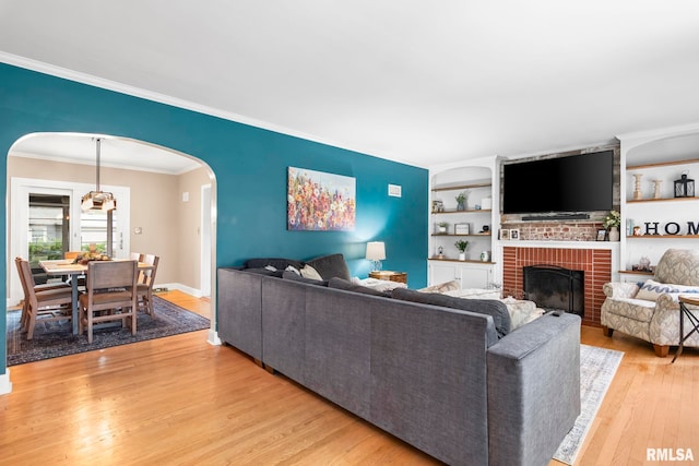 living room with light wood finished floors, arched walkways, crown molding, a fireplace, and built in shelves