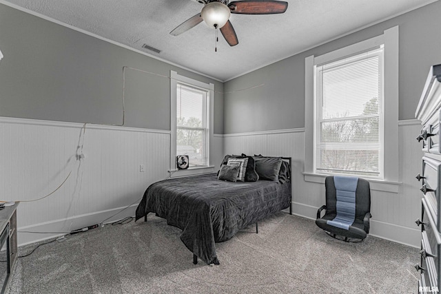 bedroom featuring a textured ceiling, visible vents, carpet, a ceiling fan, and wainscoting