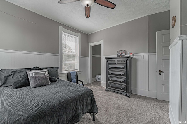 carpeted bedroom with a ceiling fan, ornamental molding, a wainscoted wall, and a textured ceiling