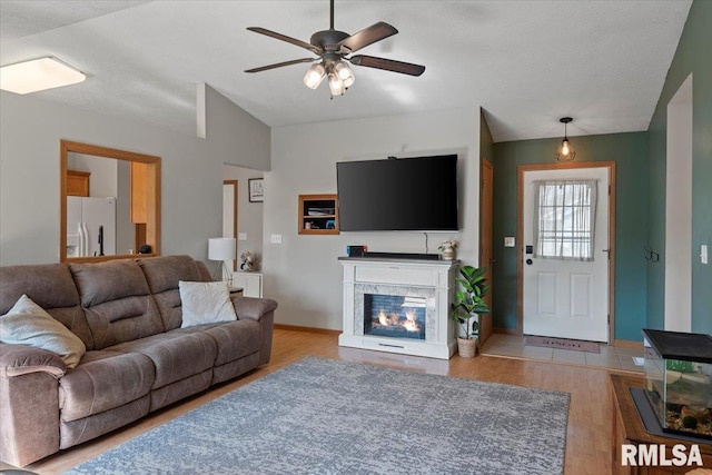 living area featuring a glass covered fireplace, light wood-style flooring, a ceiling fan, baseboards, and vaulted ceiling