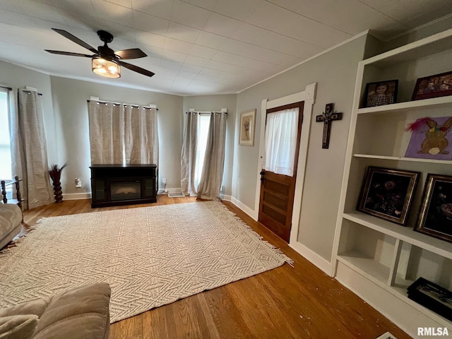 living room featuring crown molding, a glass covered fireplace, wood finished floors, ceiling fan, and built in shelves