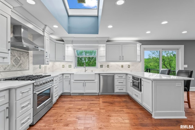 kitchen with a sink, a peninsula, stainless steel appliances, wall chimney range hood, and a breakfast bar