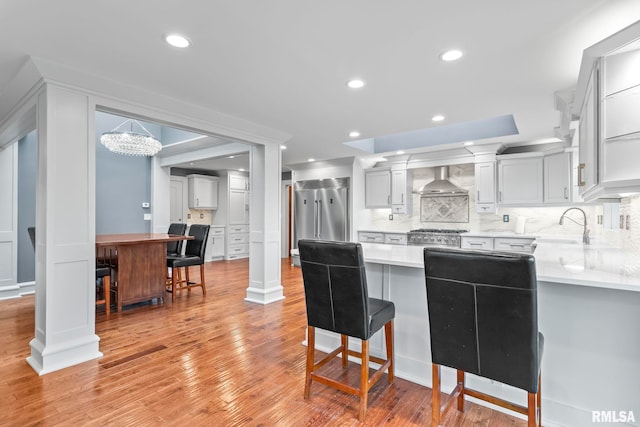 kitchen featuring built in fridge, a peninsula, wall chimney exhaust hood, decorative backsplash, and light wood-type flooring