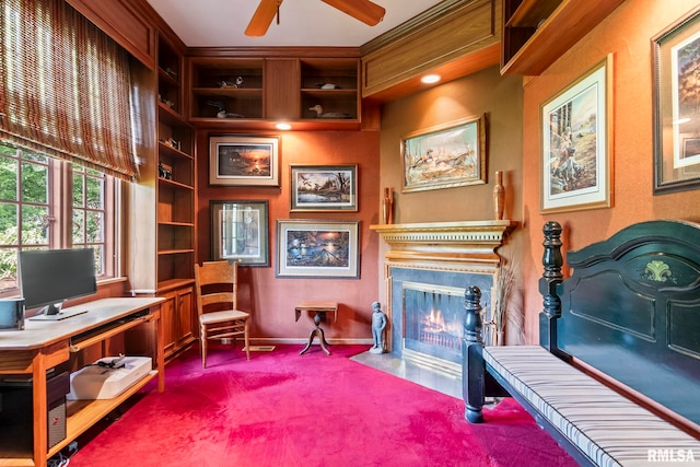 office with a fireplace with flush hearth, dark colored carpet, built in shelves, baseboards, and a ceiling fan
