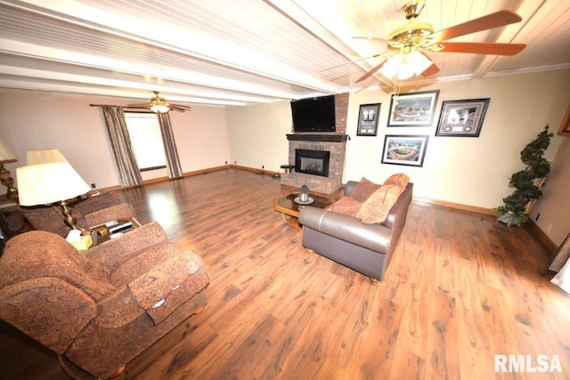 living room with ceiling fan, a brick fireplace, beamed ceiling, wood-type flooring, and ornamental molding