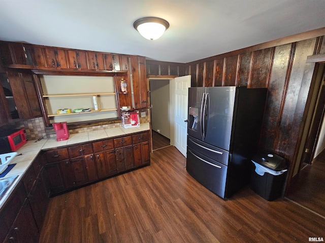 kitchen with wooden walls, dark brown cabinets, tile counters, black fridge, and dark hardwood / wood-style floors