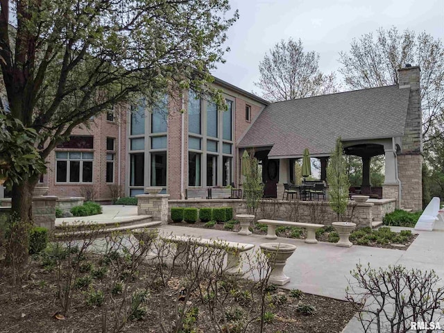 view of front of house with a patio, brick siding, roof with shingles, and a chimney