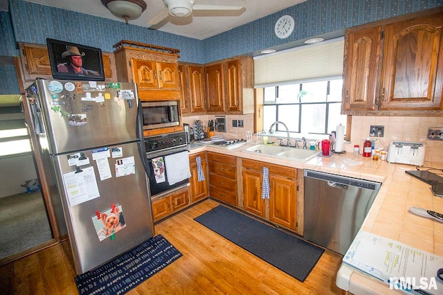 kitchen with ceiling fan, sink, appliances with stainless steel finishes, tile counters, and light wood-type flooring