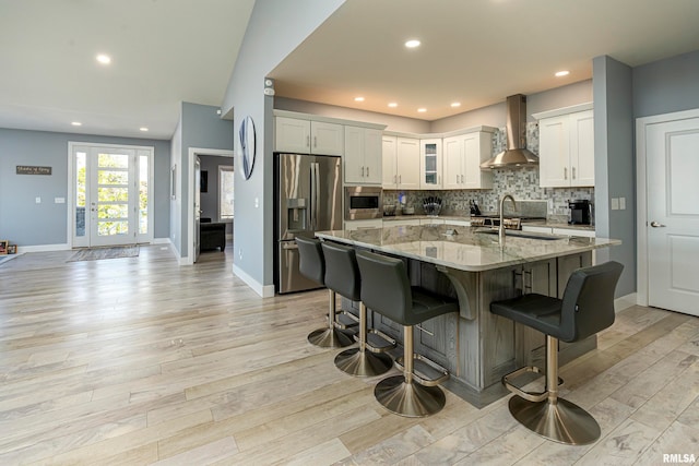 kitchen with white cabinetry, wall chimney range hood, light stone counters, an island with sink, and appliances with stainless steel finishes