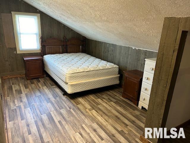 bedroom with a textured ceiling, wood walls, dark wood-type flooring, and vaulted ceiling