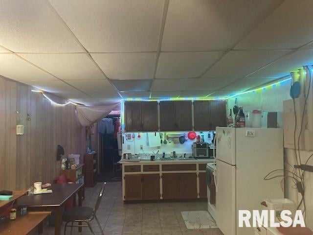 kitchen with sink, white refrigerator, a paneled ceiling, wooden walls, and light tile patterned flooring