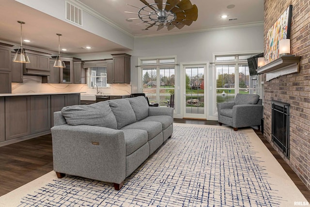 living room featuring a brick fireplace, ceiling fan, crown molding, dark wood-type flooring, and sink