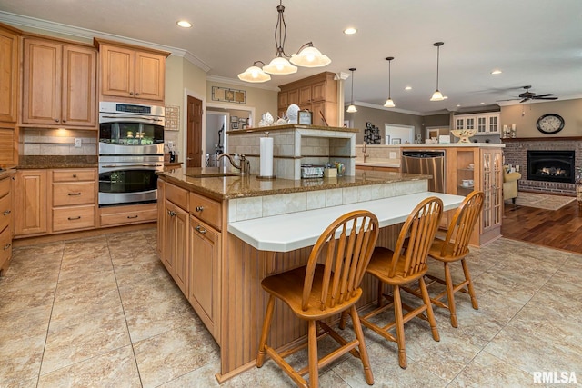 kitchen featuring hanging light fixtures, stainless steel appliances, a brick fireplace, an island with sink, and ornamental molding