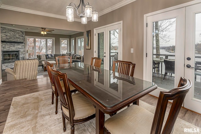 dining area with french doors, ceiling fan with notable chandelier, a stone fireplace, crown molding, and light hardwood / wood-style floors