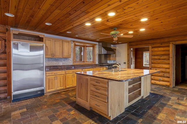 kitchen with wall chimney range hood, wood ceiling, sink, built in refrigerator, and a center island with sink