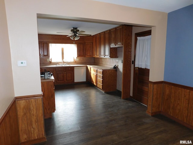 kitchen with wood walls, dark wood-type flooring, white dishwasher, sink, and ceiling fan