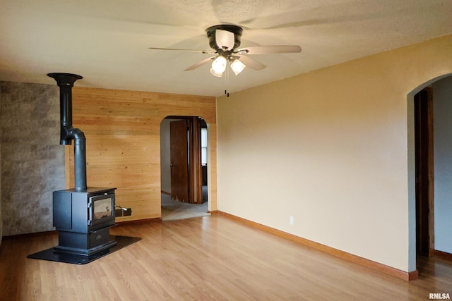 unfurnished living room with wooden walls, wood-type flooring, a wood stove, ceiling fan, and a textured ceiling