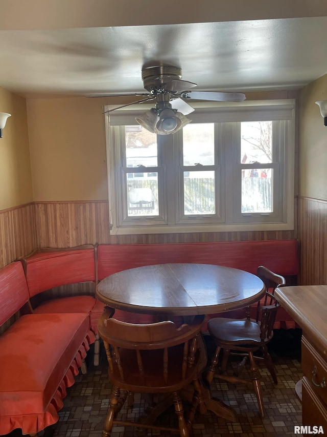 dining room featuring a wainscoted wall, ceiling fan, wooden walls, and brick floor