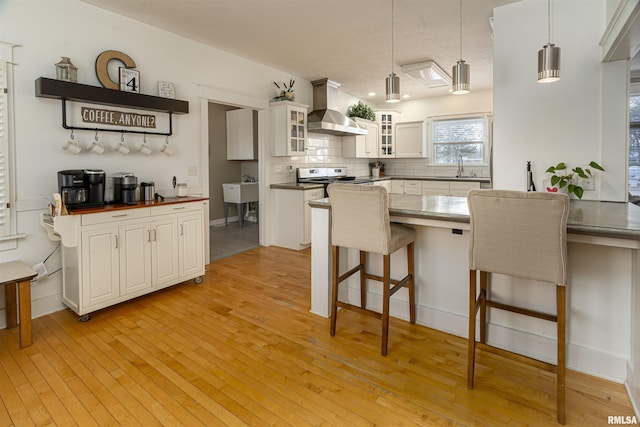 kitchen with dark countertops, hanging light fixtures, glass insert cabinets, white cabinetry, and wall chimney range hood