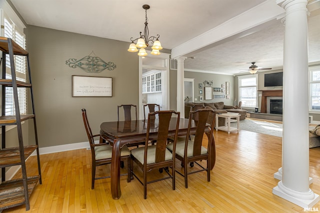 dining space featuring a fireplace with raised hearth, ceiling fan with notable chandelier, baseboards, light wood finished floors, and decorative columns