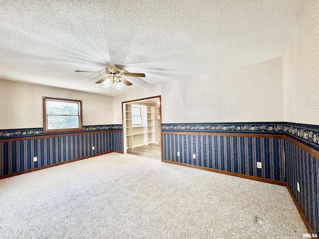 empty room with wallpapered walls, a ceiling fan, a wainscoted wall, carpet, and a textured ceiling